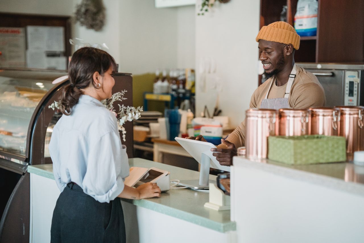 Home A barista and customer enjoy a friendly interaction at a café counter with a warm ambiance.